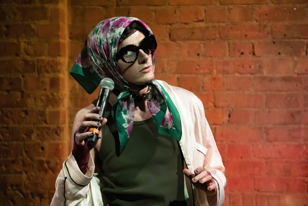 An actor wearing sunglasses and a headscarf holds a microphone.